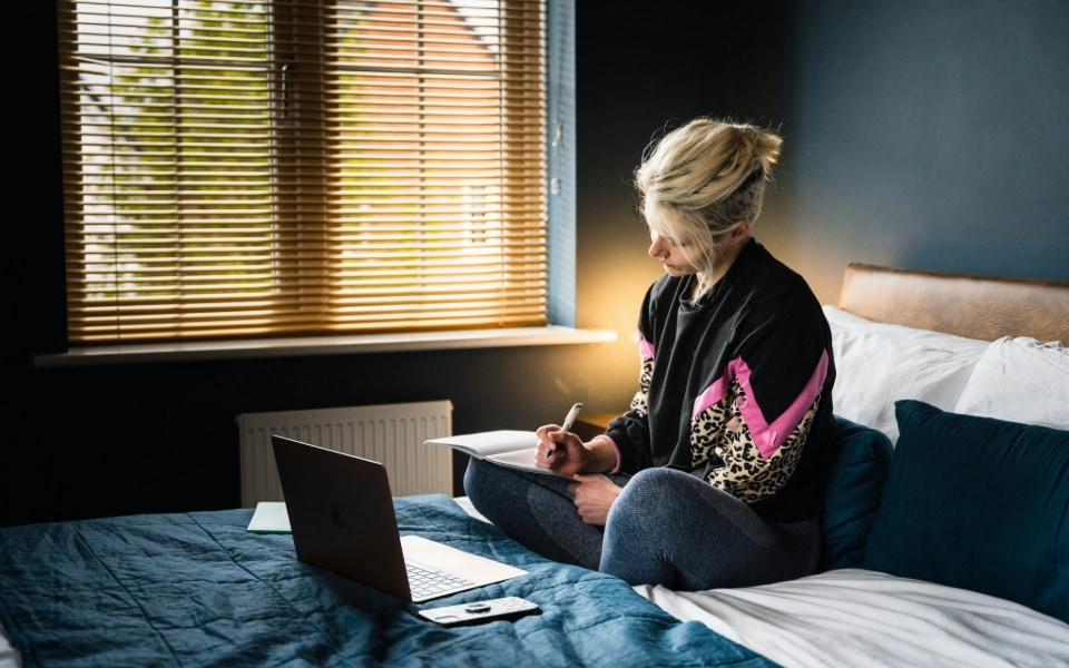 Eine Frau schreibt auf einem Bett in einem Raum mit natürlichem Licht, das durch Jalousien fällt, mit aufgeklapptem Laptop in ein Notizbuch.