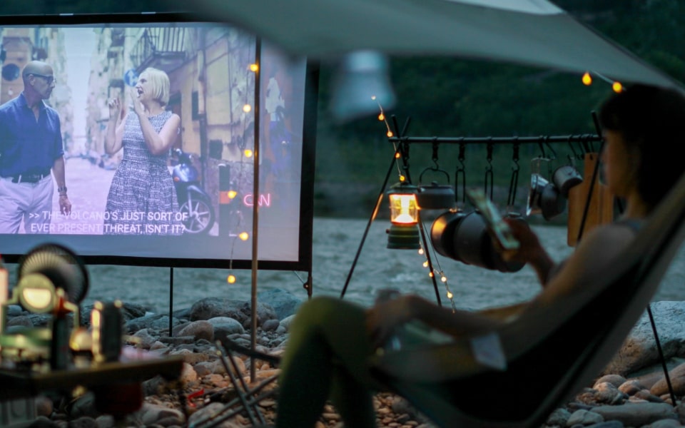 Eine Person entspannt sich in einem Stuhl unter einem Baldachin bei einem Freiluftkino am Flussufer, mit Laternen im Hintergrund.