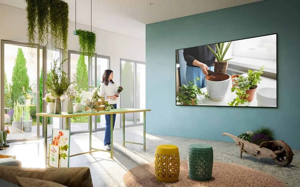 Was bedeutet Smart-TV? Beste Unterhaltung für diese Frau in einem hellen, mit Pflanzen gefüllten Wohnraum mit einem wandmontierten LG-Fernseher, der die Benutzeroberfläche des webOS-Homescreens abbildet. 