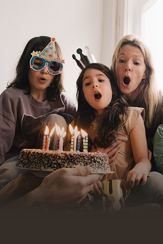 Billede af to voksne kvinder og en ung pige, som er iført fødselsdagshatte, mens de blæser lyd ud på kagen.