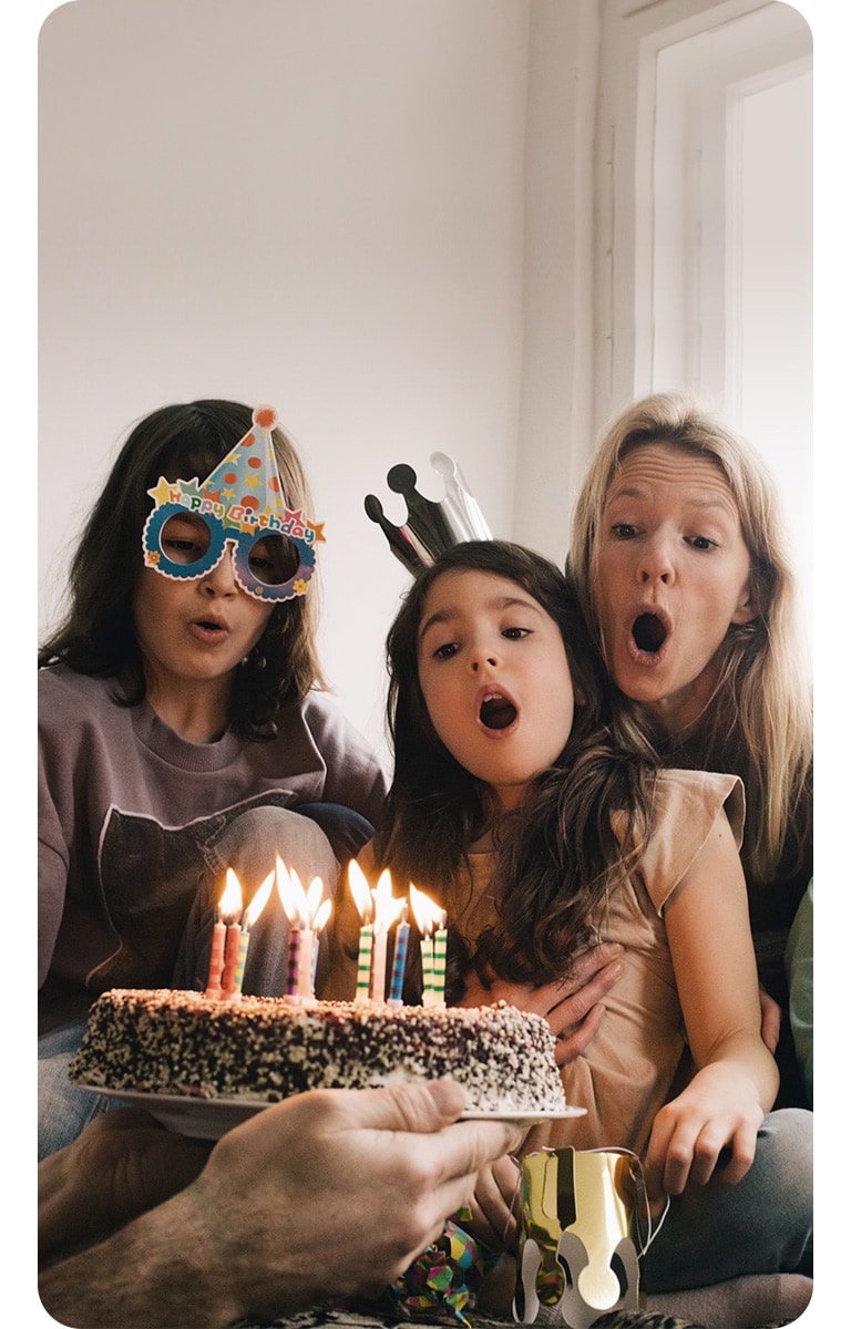 Billede af to voksne kvinder og en ung pige, som er iført fødselsdagshatte, mens de blæser lyd ud på kagen.