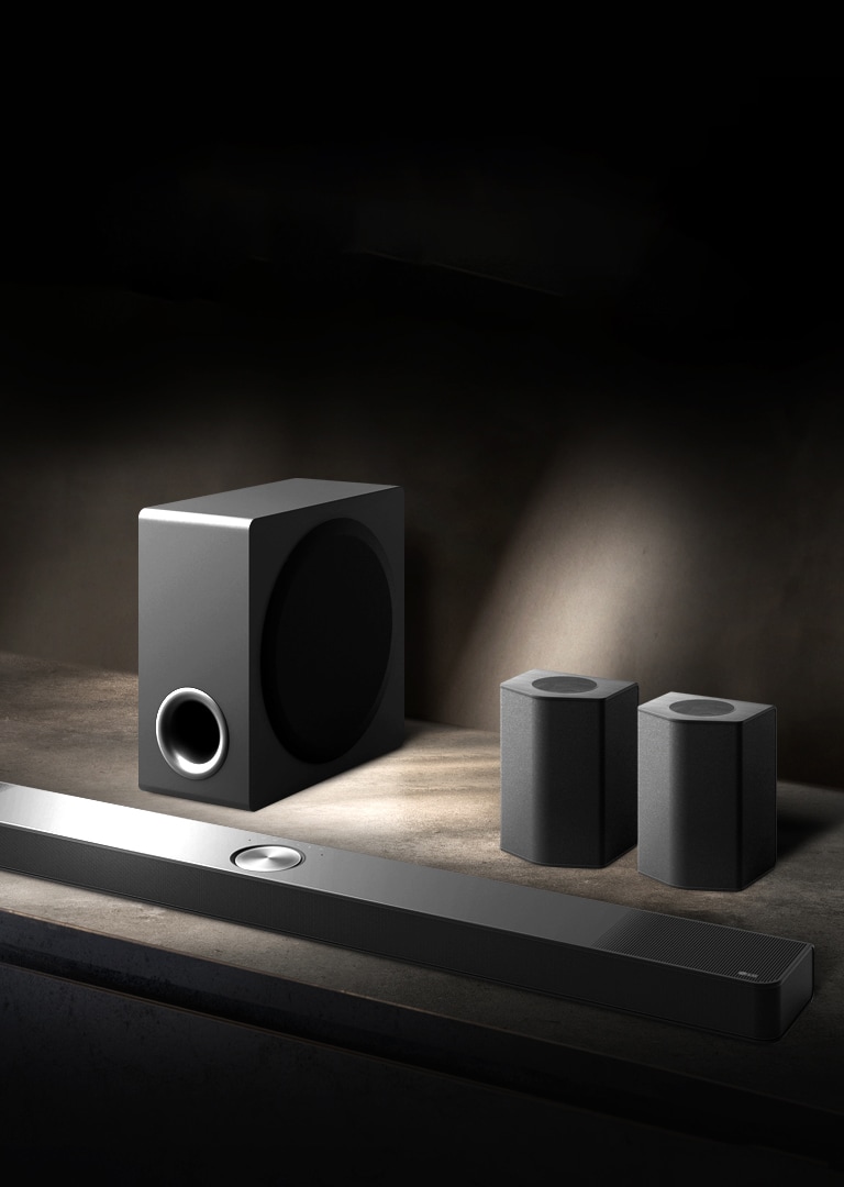 La LG Soundbar, les enceintes arrière et le caisson de basse installés en biais sur une étagère en bois marron dans une pièce obscure, avec une lumière qui n’éclaire que le système audio.