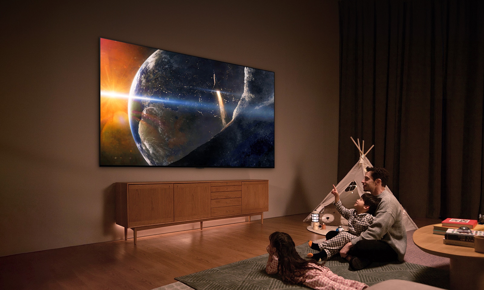 A family sat on the floor of a low-lit living room by a small table, looking up at an LG TV mounted on the wall showing the Earth from space.