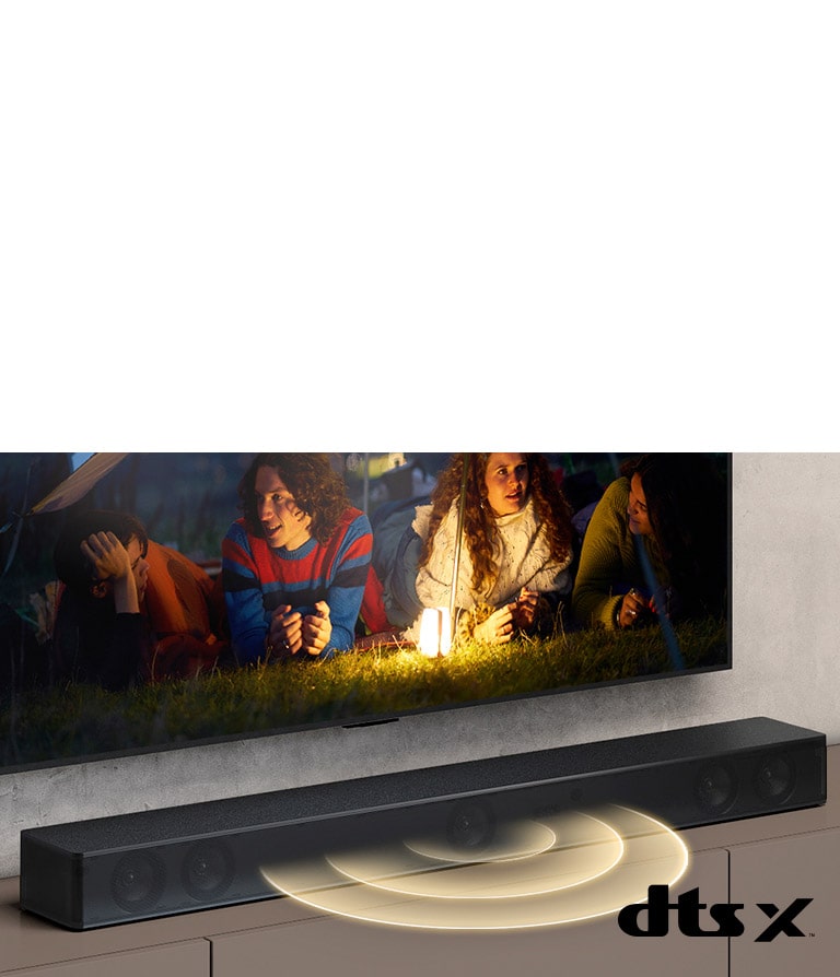 LG TV is on the wall, on the screen it shows 2 couples lying on the grass. In front of them, there is a lamp. LG Sound bar is below LG TV. Sound graphic is coming out from the front of the sound bar. DTS Virtual:X logo is shown on the bottom right corner of image.