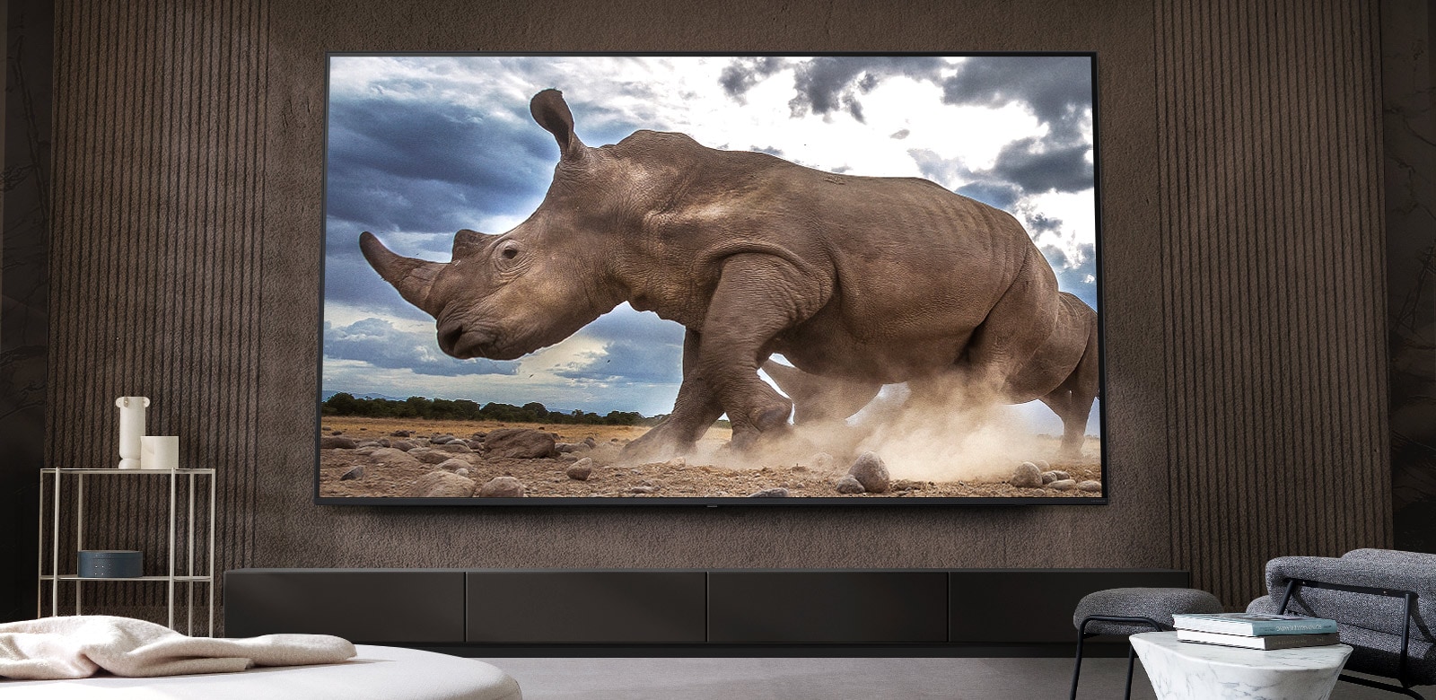 Un rinoceronte en un entorno de safari se muestra en un televisor LG ultra grande, montado en la pared marrón de una sala de estar rodeada de muebles modulares de color crema.