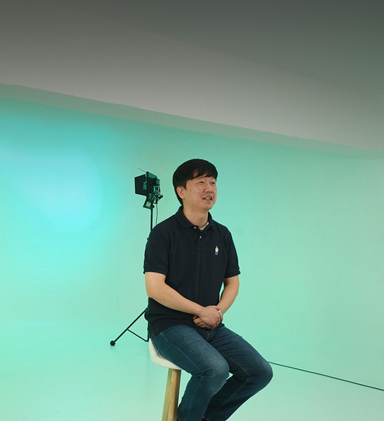 LG CAE Team Member wearing black short-sleeved shirt is sitting on a chair with green background.