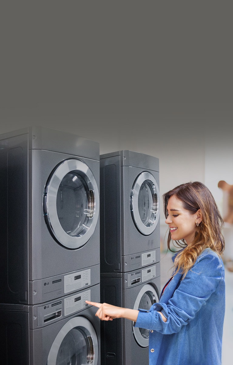 A smiling woman is interacting with a row of modern dark gray LG Commercial Laundry in a bright laundry room, demonstrating the convenience and ease of maintenance of the efficient system.