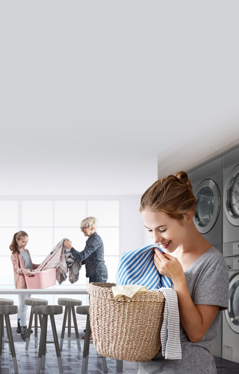 A modern laundry room with multiple LG Commercial Laundry features a woman smelling freshly laundered clothes from a basket and other customers interacting in the background.