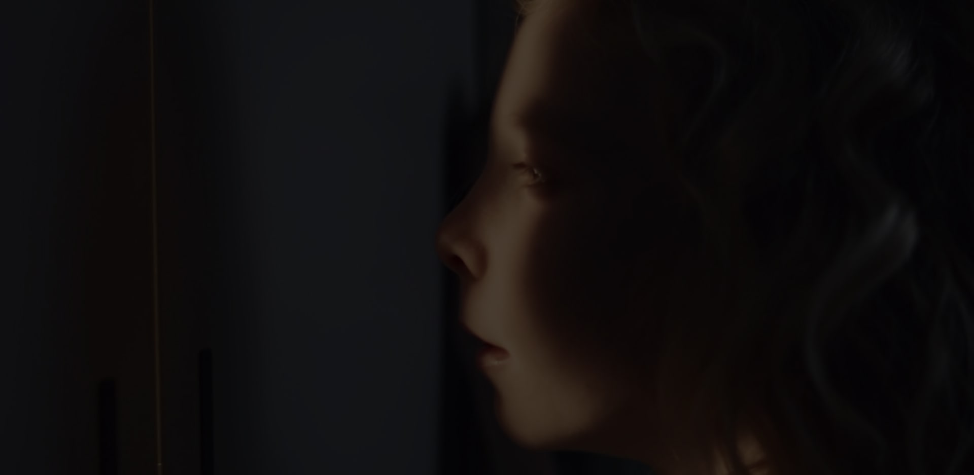 A close-up shot of a girls face, partially in shadow, partially illuminated as she peers out from a hiding place.