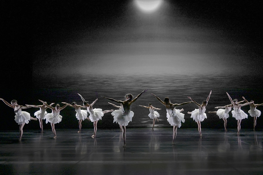 Ballerinas pose with arms and legs raised on a dark stage with a backdrop of moonlight on a lake.