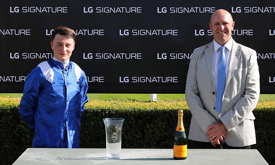 a winning jockey from the Qatar Goodwood Festival is presented with a glass trophy and a bottle of champagne in front of an LG SIGNATURE-branded sign.