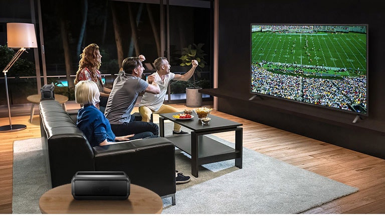 Μια παρέα παρακολουθεί αθλητικά παιχνίδια στην τηλεόραση στο σαλόνι, με τα ηχεία Bluetooth πίσω τους.