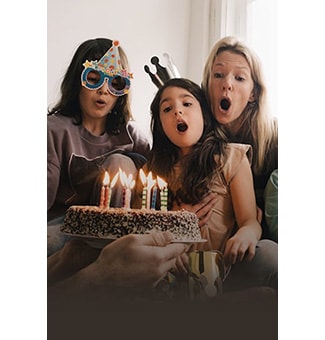 'Μια εικόνα δύο γυναικών και ενός κοριτσιού που φοράει ένα καπελάκι γενεθλίων και φυσάει τα κεριά σε μια τούρτα.