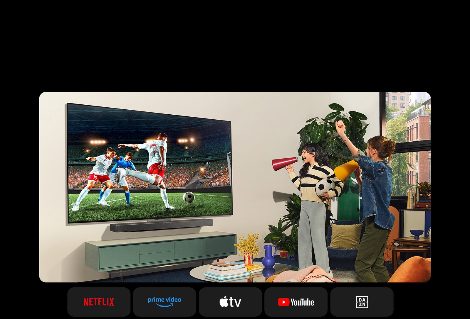 Két nő focimeccset néz egy hangulatos nappaliban. Az egyik nő egy focilabdát tart a kezében, és mindketten szurkolnak a csapatuknak sárga és piros hangszórókkal. Lent a következő logók láthatók: Netflix, Amazon Prime Video, Apple TV, Youtube, ESPN és DAZN.