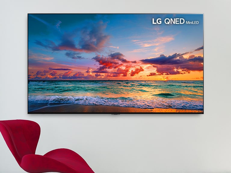 Egy falra rögzített nagyképernyős TV, előtte egy piros szék. A képernyőn egy tengerpart látható, amelyek finoman mosnak a hullámok. Balról jobbra görgetve láthatóvá válik a kép hagyományos LCD képernyőn, valamint egy LG QNED MiniLED képernyőn, mutatva a színkülönbséget.