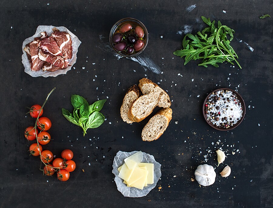 Assortimento di ingredienti freschi, tra cui carne, olive, pomodori, aglio, pane, erbe e spezie su una superficie nera