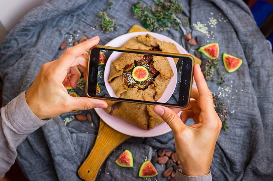 Un paio di mani tengono uno smartphone sopra un tortino di fichi freschi mentre viene scattata una foto del piatto.