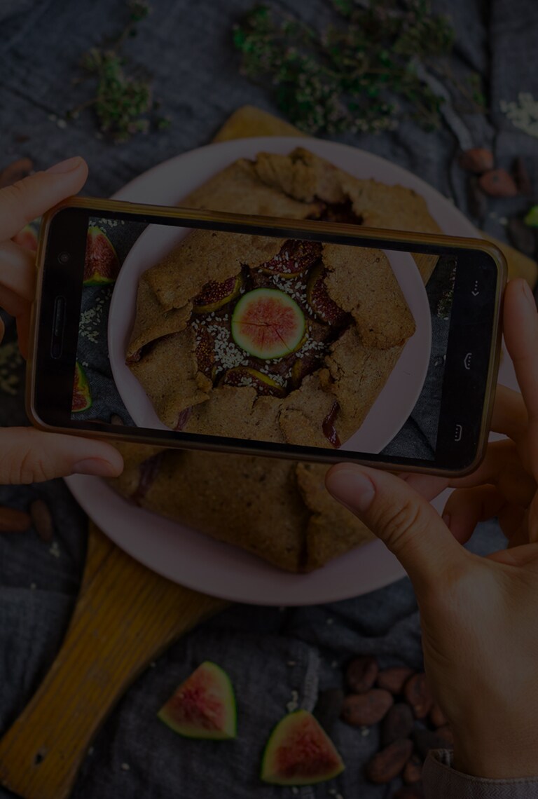 Un paio di mani tengono uno smartphone sopra un tortino di fichi mentre viene scattata una foto. La scritta 'Siamo ciò che mangiamo' è sovrapposta all'immagine.
