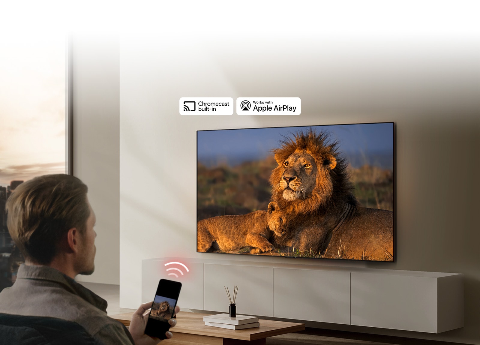 An LG TV mounted on a wall in a living room, displaying a lion and lion cub. A man sits in the foreground with a smartphone in his hand displaying the same image of lions. A graphic of three neon red curved bars is displayed just above the smartphone pointing toward the TV.