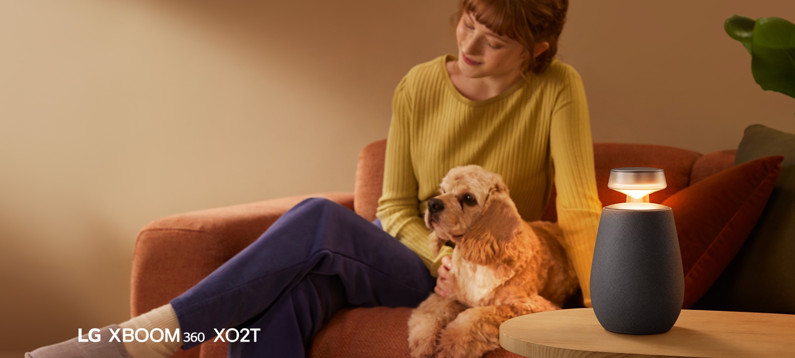 Sieviete sēž uz dīvāna ar suni, klausoties mūziku ar LG XBOOM 360 XO2T.
