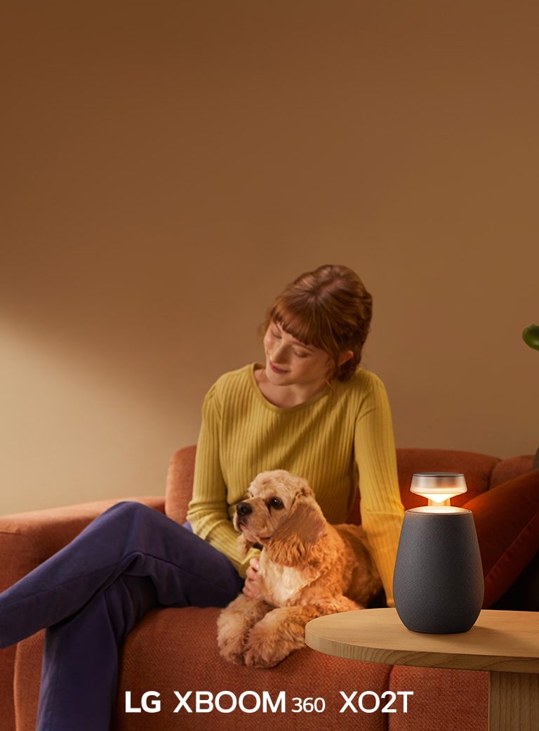 Sieviete sēž uz dīvāna ar suni, klausoties mūziku ar LG XBOOM 360 XO2T.