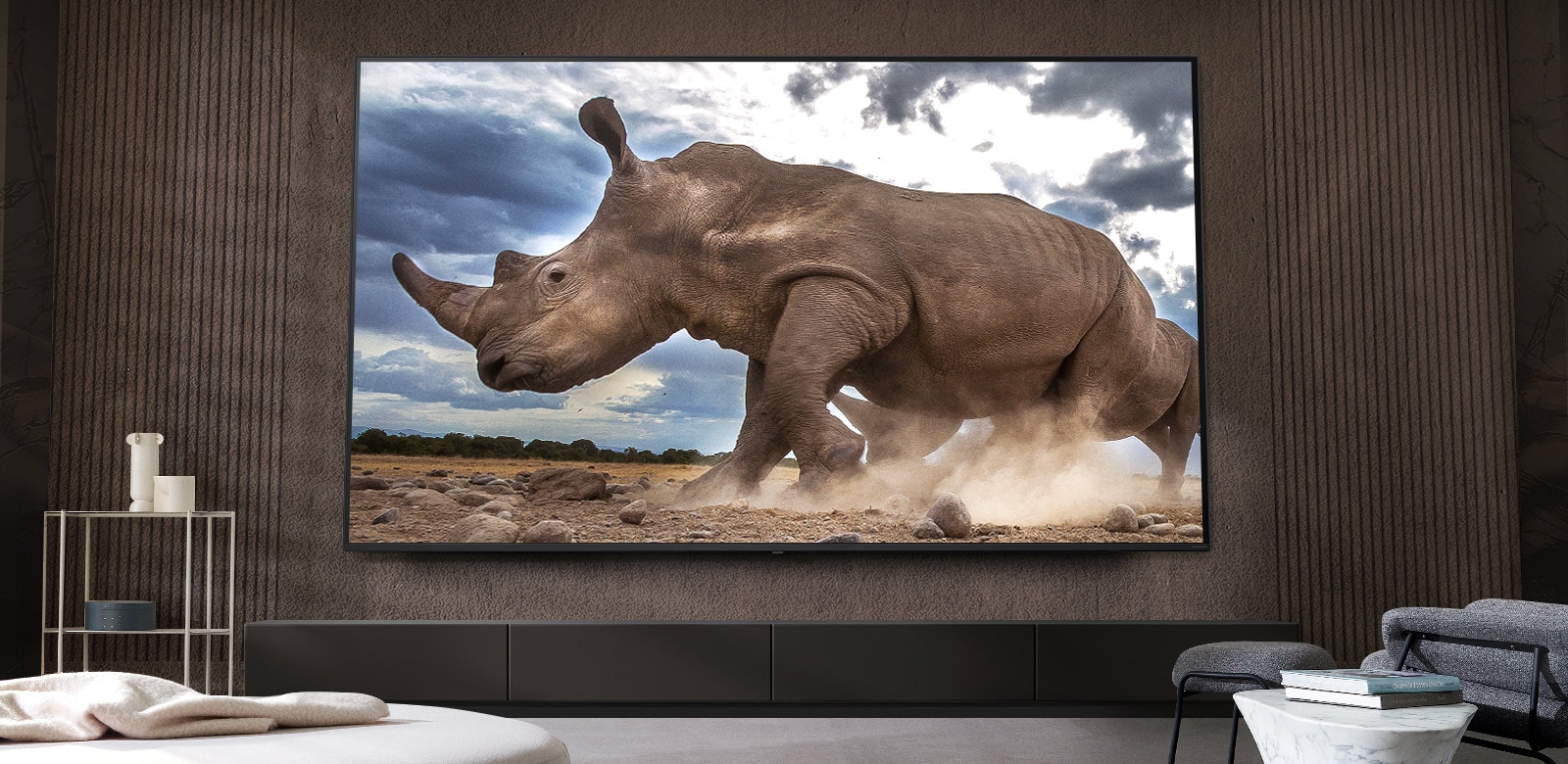 A rhinoceros in a safari setting is shown on an Ultra Big LG TV, mounted on the brown wall of a living room surrounded by cream-colored modular furniture.