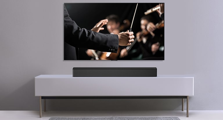 A TV is shown on a gray wall and LG Soundbar below it on a gray shelf. TV shows a conductor conducting an orchestra. 