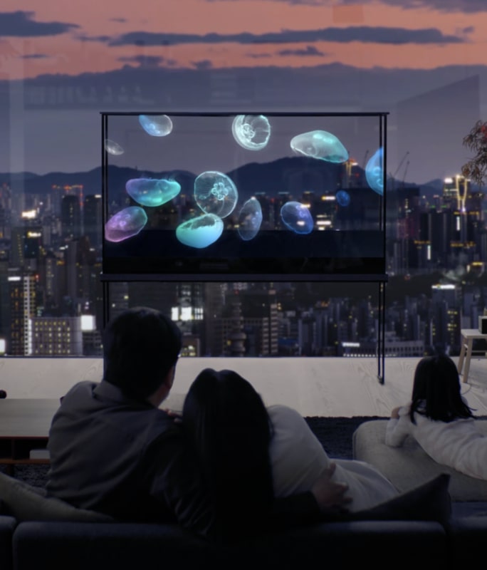 A family enjoys an LG Oled TV with a jellyfish floating on it, while the cityscape shines through the transparent screen. 