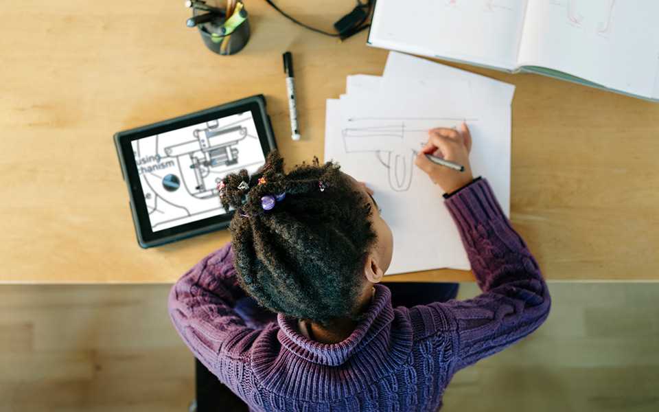 A child sitting at a desk, drawing on a piece of paper with an iPad on her left.