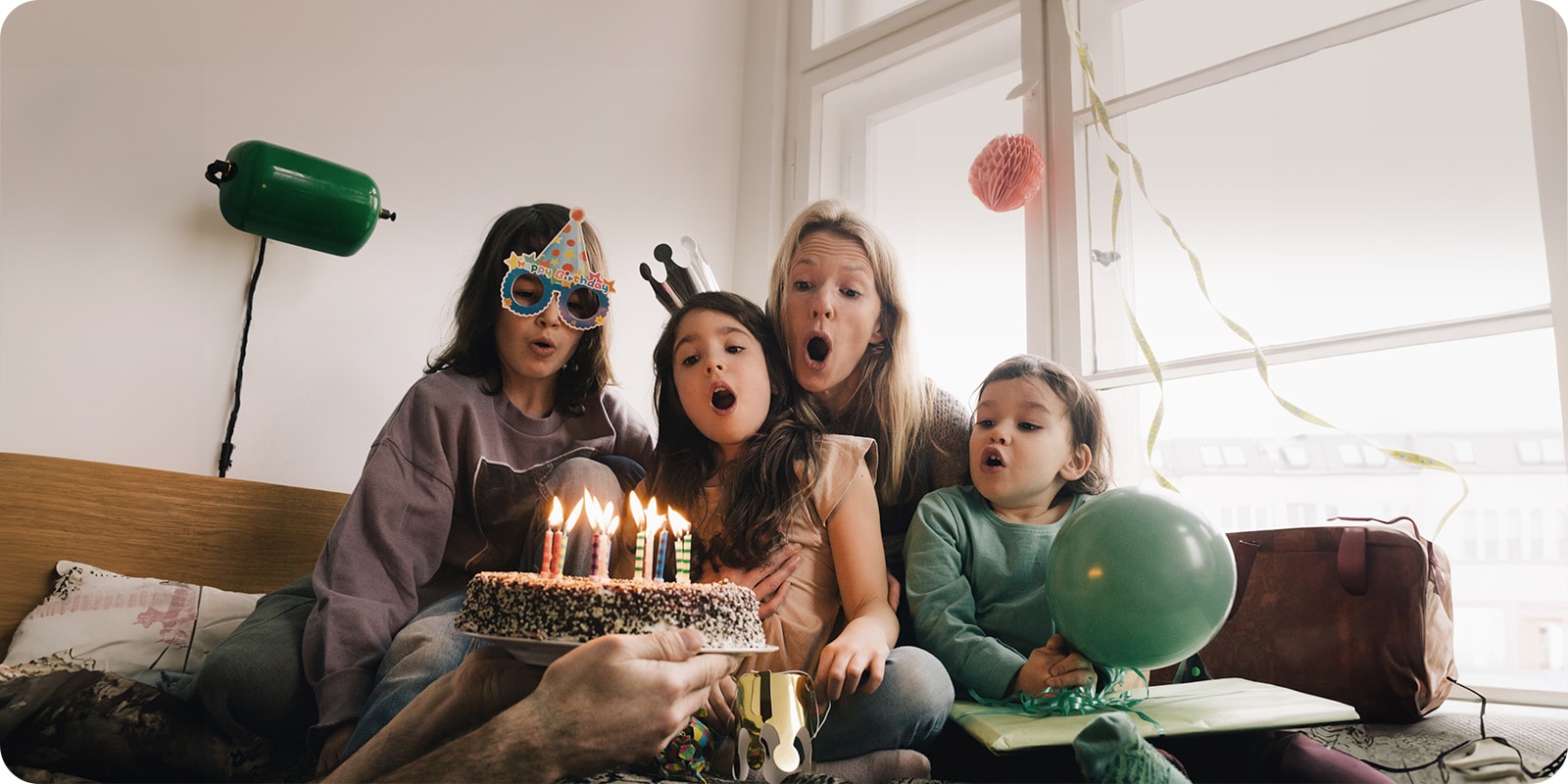 Bilde av to voksne kvinner og en ung jente med bursdagshatt på hodet som blåser ut lysene på kaken.