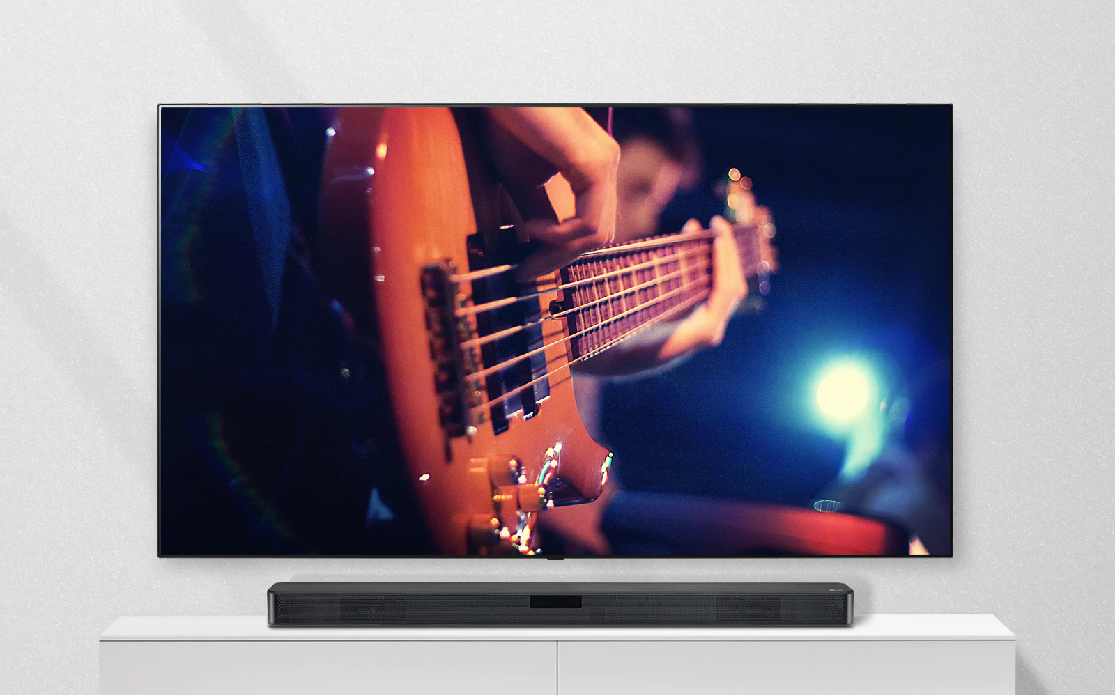 The TV is attached to the wall, and the sound bar is on a white shelf. TV showing a man plays guitar.