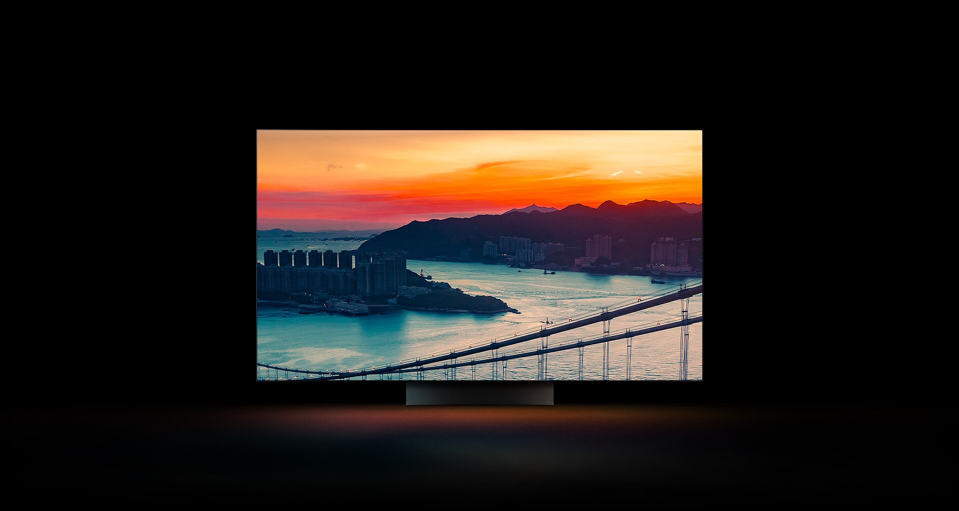 Prikaz grada na obali iz vazduha sa saobraćajem koji se kreće preko mosta dok sunce zalazi pojavljuje se preko celog ekrana. Isečen je i prikazan na ekranu LG OLED televizora sa neizmenjenim originalnim bojama.