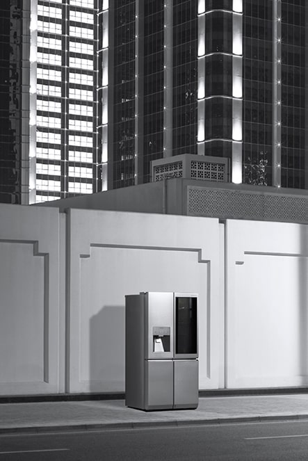 Холодильник LG SIGNATURE стоит на ночной улице со зданиями, из окон которых исходит свет