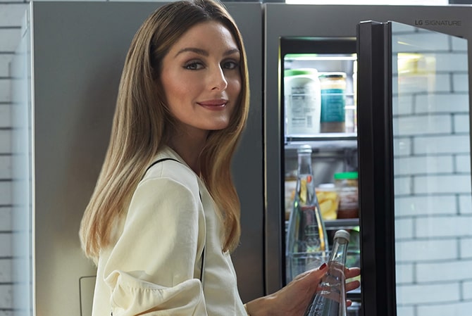 Оливия Палермо открывает стеклянную панель instaview холодильника LG SIGNATURE: на полках видны готовые закуски.