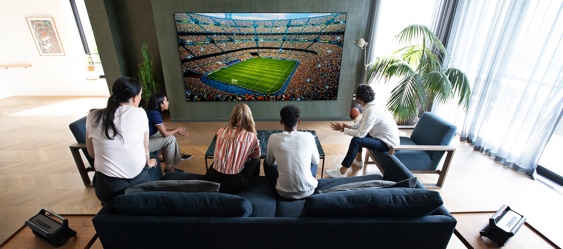 A group of friends watching a sports game on TV in the living room with 2 same Bluetooth speakers at rear speaker position