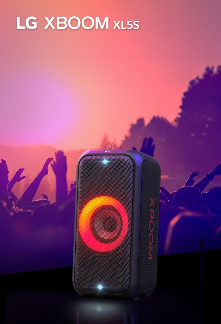 LG XBOOM XL5S umiestnený na pódiu so zapnutým červeno-oranžovým osvetlením. Za pódiom si ľudia užívajú hudbu.