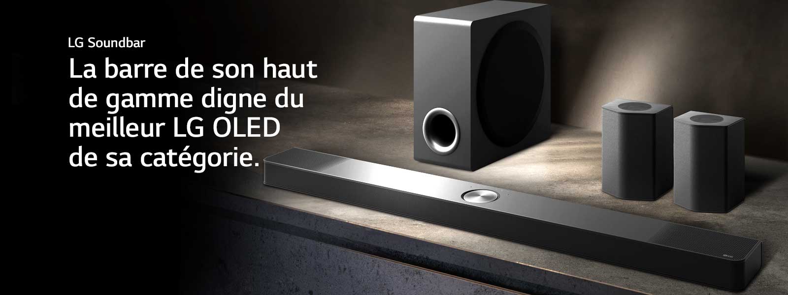 La LG Soundbar, les haut-parleurs arrière et le caisson de basse sont installés en biais sur une étagère en bois marron dans une pièce obscure, avec une lumière qui n’éclaire que le système audio.