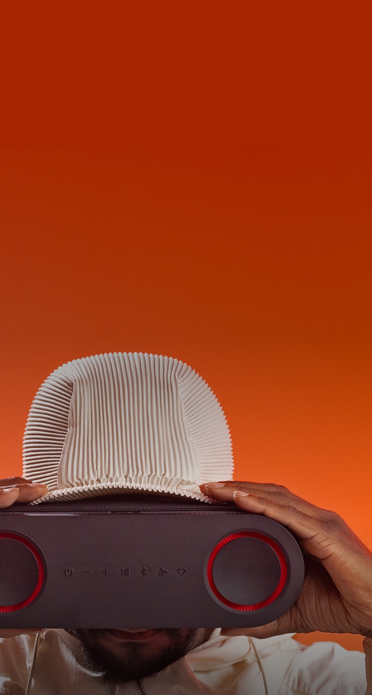 Photograph of will.i.am against an orange background faces the camera and holds an LG xboom speaker in front of his face like goggles.