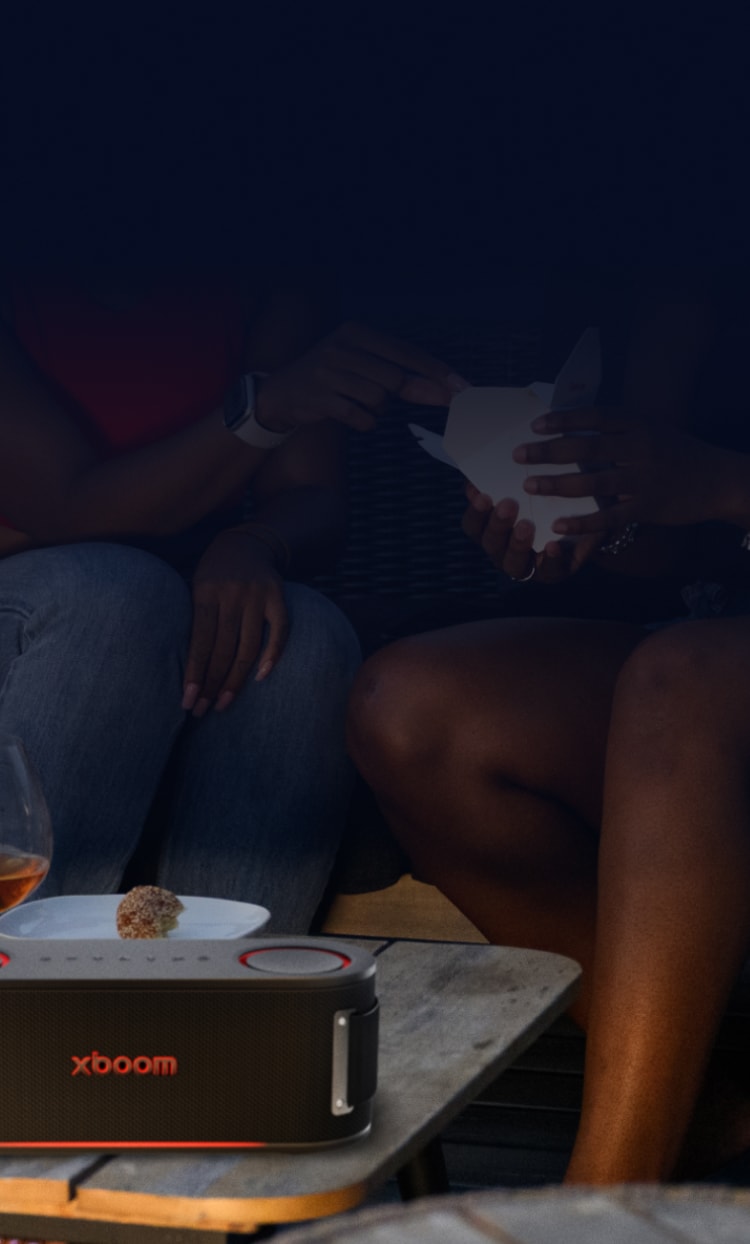 Smartphone displaying the LG ThinQ app interface with two people relaxing on a couch in the background, and an LG xboom speaker in the foreground.