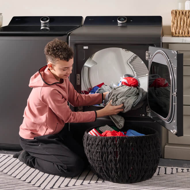 A teenager is taking clothes out of a front-load dryer and putting them into a laundry basket on the floor of a laundry room.