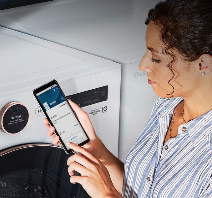 A woman stands in front of her top-load washer using the ThinQ app on her phone to control the washer.