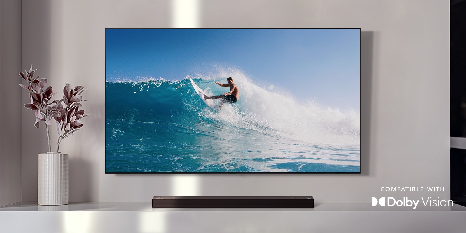 TV is on the wall. TV shows a man surfing on big wave. LG Soundbar is right below TV on a white shelf. There is a vase with a flower right next to the soundbar. (play the video)