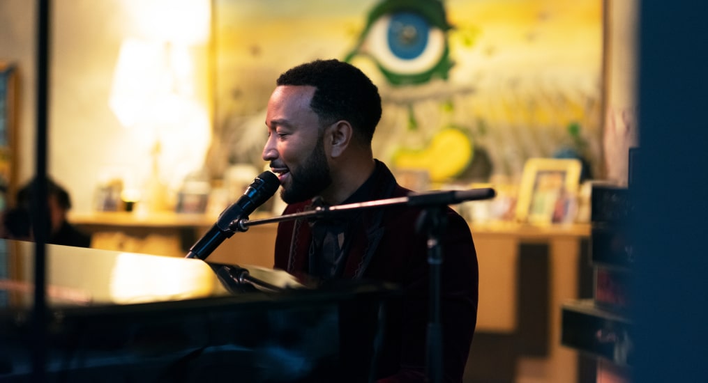 A side shot of John Legend as he sings into the mic while playing the piano.