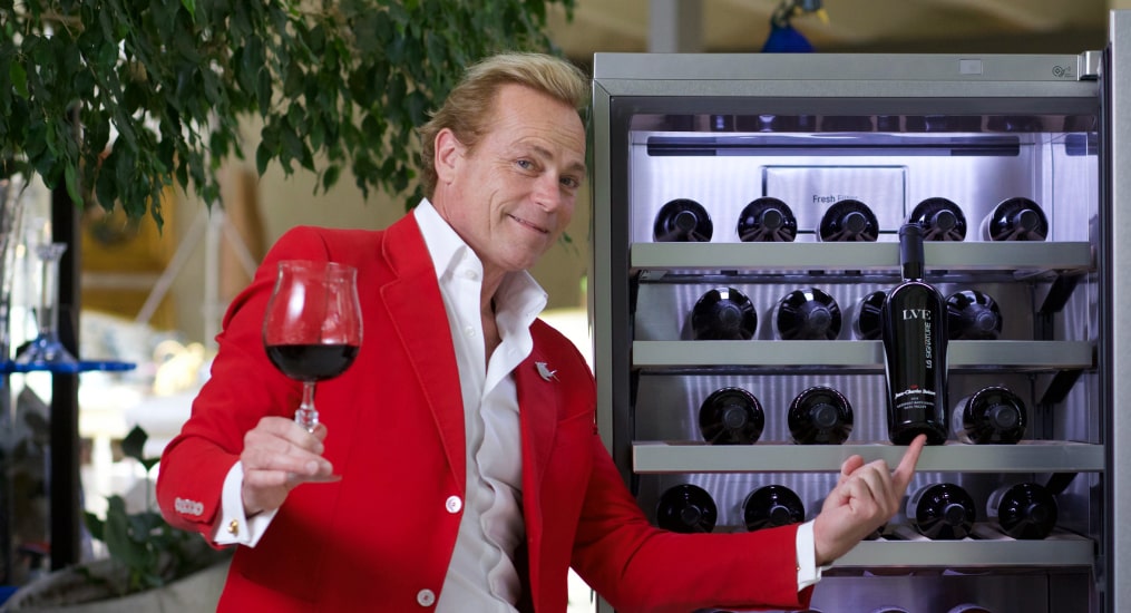 Jean-Charles Boisset look into the camera playfully while holding a glass of wine in one hand, a bottle in the other. He is in a red suit and next to a fully stocked wine cellar.