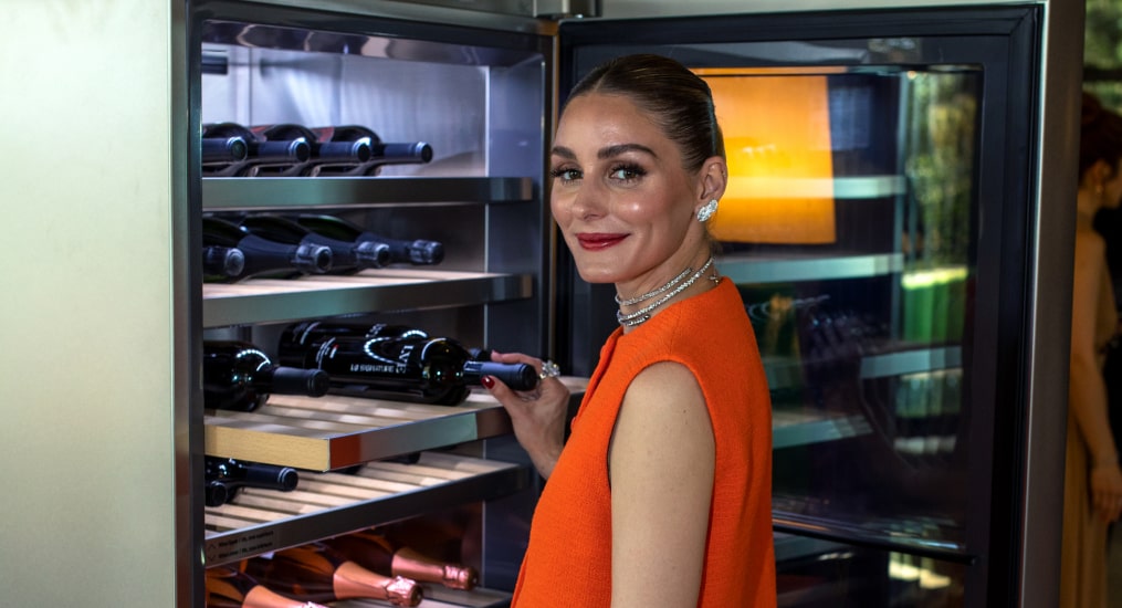 Also clad in red, Olivia Palermo stands in front of an open wine cellar. She holds the neck of a wine bottle as if to take one out, with a smile toward the camera.