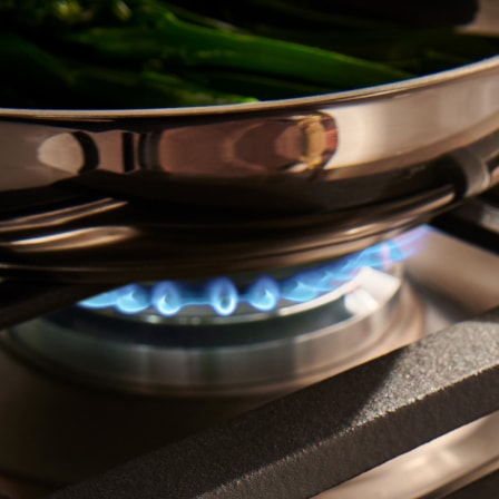 Close-up view of a gas burner with blue flames, cooking food in a stainless steel pan. The word 'Gas' is displayed in the bottom corner.