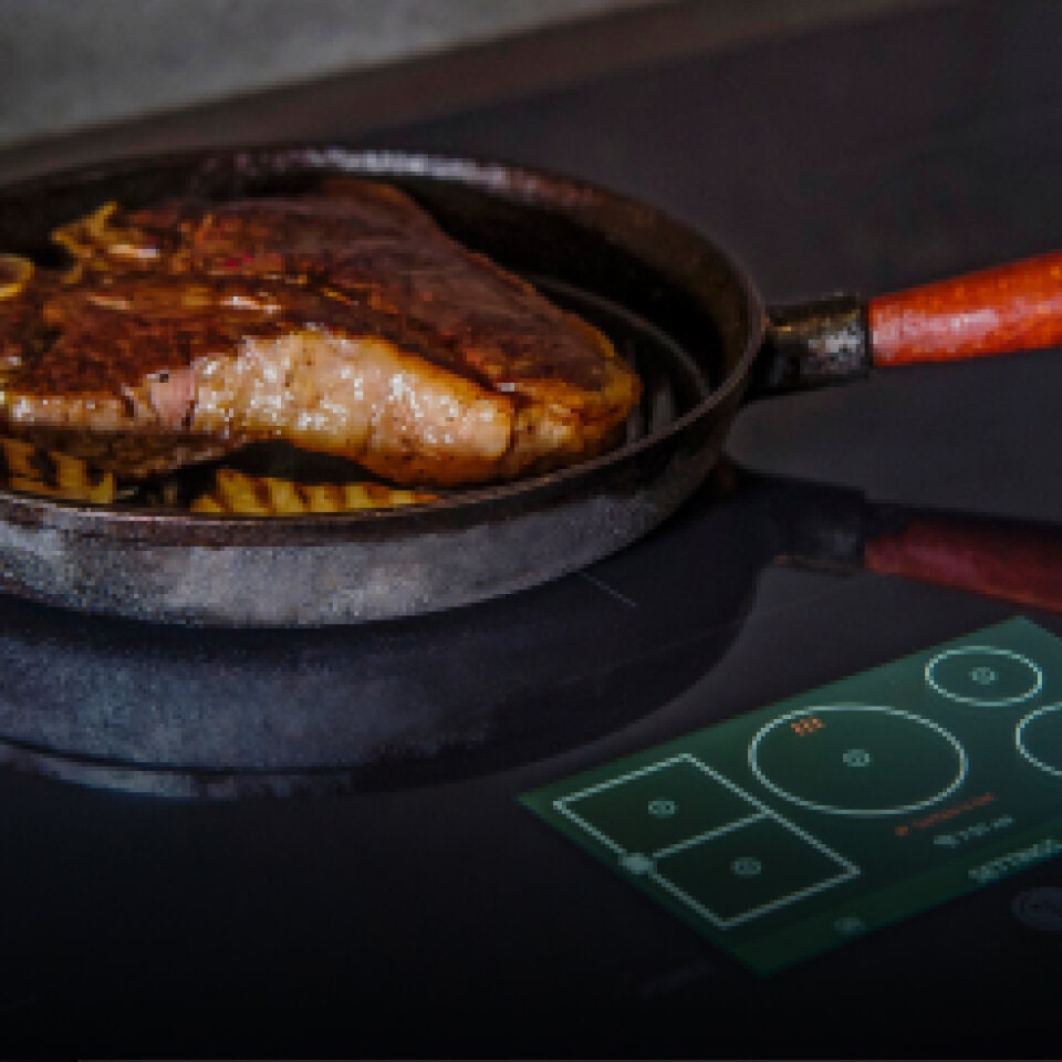 A steak searing in a hot cast iron pan on an induction cooktop with temperature controls visible. The word 'Induction' is displayed in the bottom corner.