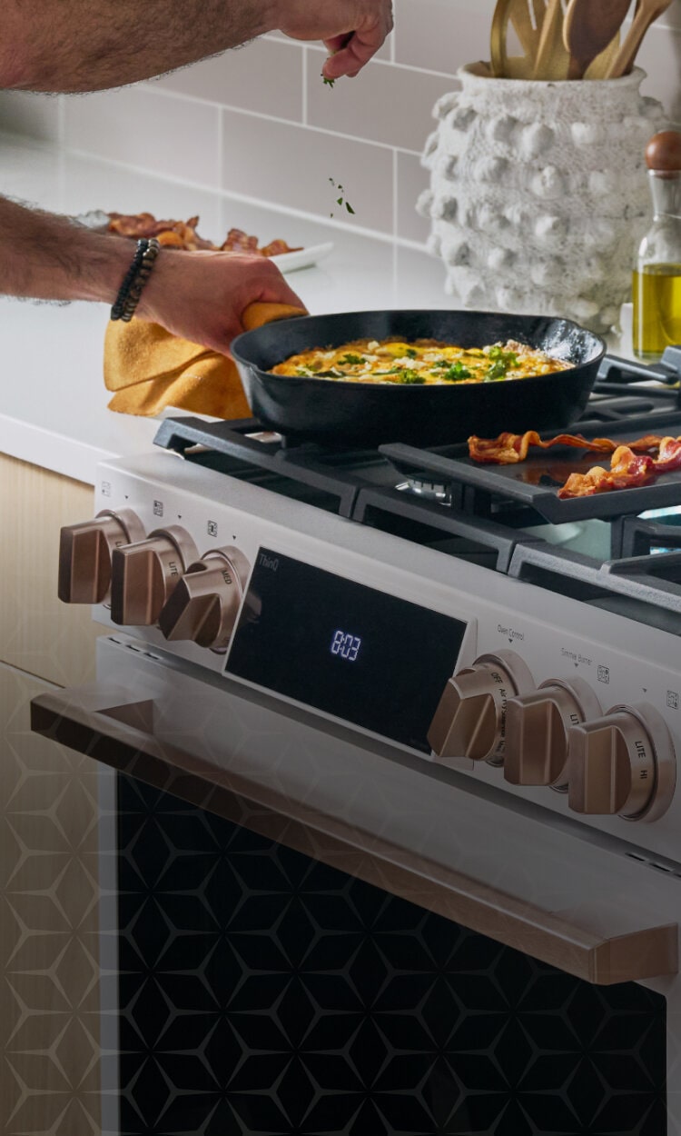 Person cooking breakfast on an LG Studio gas range, frying bacon and preparing a dish in a cast-iron pan.