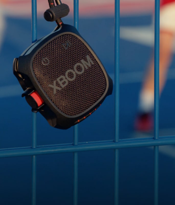 A close-up of a portable XBOOM Go Bluetooth speaker hanging on a blue fence, with a blurred outdoor background.