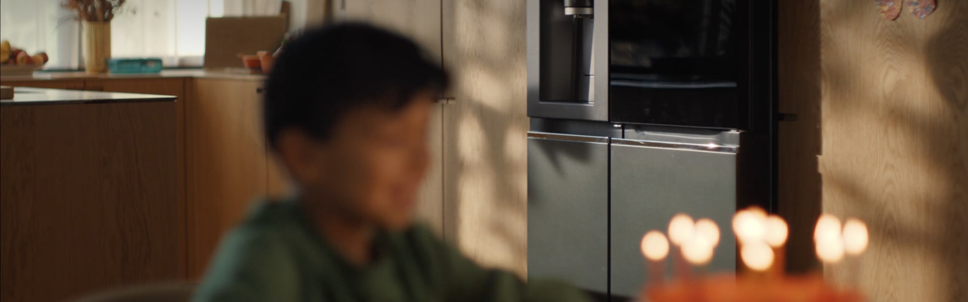 The child's face is faintly visible in front of the candlelit cake, and the LG InstaView refrigerator is clearly visible in the background.